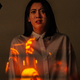 A dark-haired woman wearing a white button down shirt stands with a look of fear on her face as disturbing images are projected across her white shirt.